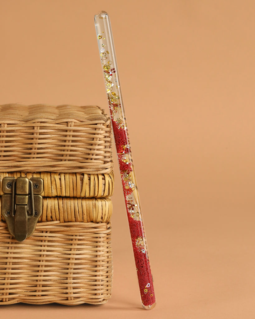 The Konges Sloejd Magical Glitter Wand - Cherry, decorated with red and gold floral designs, leans gracefully against a closed wicker basket with a metal latch on a simple beige background.