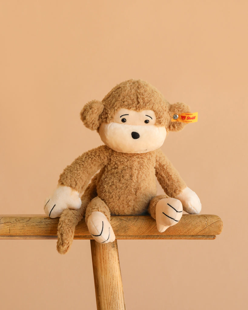 A Steiff Brownie Monkey plush with a white face, hands, and feet sits on a wooden bench against a light beige background. It features a yellow tag in its ear, making it safe for collectors and children alike.