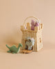 A Olli Ella Rattan Castle Bag containing plush dinosaur toys in soft pastel colors, with one toy dinosaur outside the basket on a plain beige background.