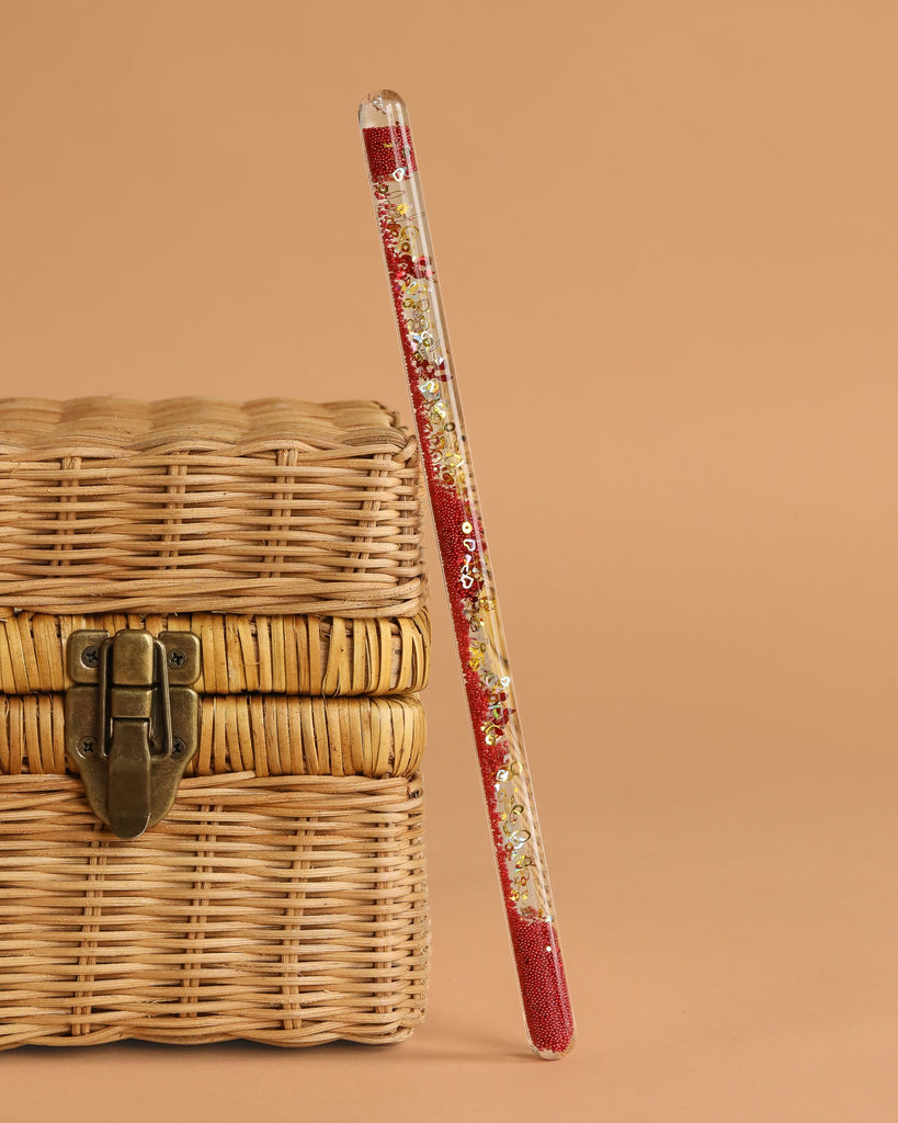 The Konges Sloejd Magical Glitter Wand - Cherry, filled with sparkling glitter, stands upright against a wicker basket with a metal latch on a beige background.