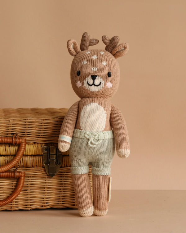 Cuddle + Kind Elliott the Fawn, a hand-knit plush deer with beige fur, white accents, antlers, and light green shorts, sits upright before a woven wicker basket on a neutral beige background.
