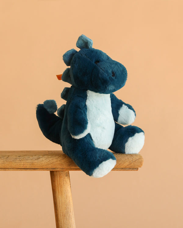 The Steiff Spott Stegosaurus Dinosaur, a blue and white plush with soft spikes, sits on the edge of a wooden bench against a plain peach background.