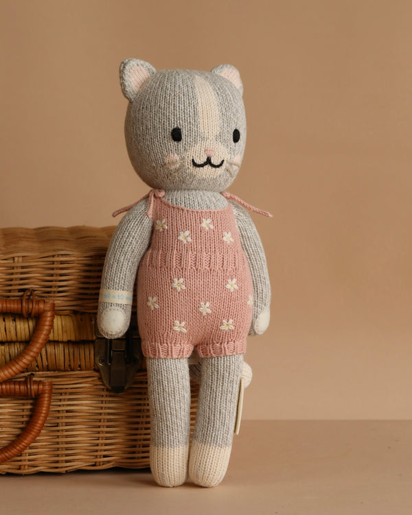 Cuddle + Kind Daisy the Kitten is a hand-knit gray cat doll in pink overalls with white flowers, smiling and sitting against a tan background beside a wicker basket. Crafted from premium cotton yarn.
