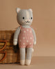 Cuddle + Kind Daisy the Kitten is a hand-knit gray cat doll in pink overalls with white flowers, smiling and sitting against a tan background beside a wicker basket. Crafted from premium cotton yarn.