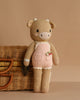 Cuddle + Kind Ava the Cow - Powder Pink is a hand-knit plush doll in a pink outfit, standing upright beside a wicker basket with a metal clasp, set against a plain beige background.