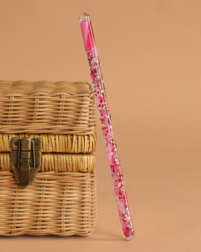 The Konges Sloejd Magical Glitter Wand - Pink Mix stands upright, leaning against a closed wicker basket with a metal latch, set against a beige background.