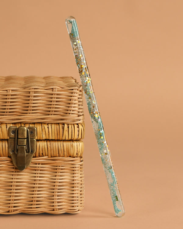 The Konges Sloejd Magical Glitter Wand - Green Mix leans against a woven wicker chest with a metal clasp, set against a plain beige background.