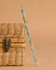 The Konges Sloejd Magical Glitter Wand - Green Mix leans against a woven wicker chest with a metal clasp, set against a plain beige background.