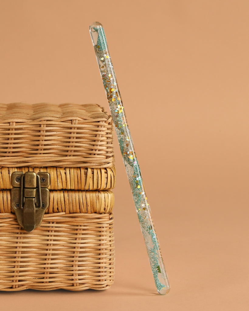 The Konges Sloejd Magical Glitter Wand - Green Mix leans against a woven wicker chest with a metal clasp, set against a plain beige background.