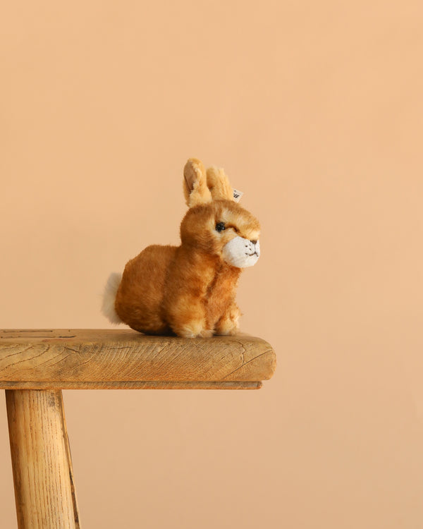 The Steiff Hoppel Rabbit, a machine washable plush bunny, sits on a wooden stool against a plain beige background, making it an ideal gift for kids.