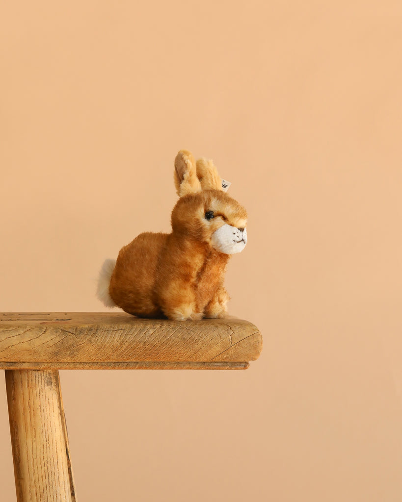 The Steiff Hoppel Rabbit, a machine washable plush bunny, sits on a wooden stool against a plain beige background, making it an ideal gift for kids.