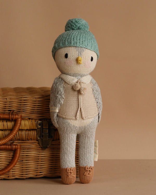 Cuddle + Kind Greyson the Penguin, a hand-knit doll with a bird-like face, wears a blue knit hat, beige sweater, and brown shoes, standing before a wicker basket on a beige background.