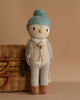 Cuddle + Kind Greyson the Penguin, a hand-knit doll with a bird-like face, wears a blue knit hat, beige sweater, and brown shoes, standing before a wicker basket on a beige background.