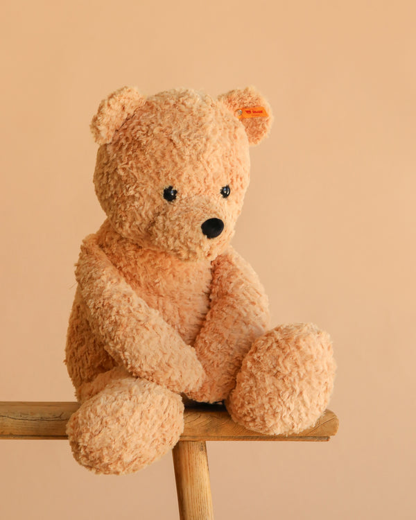 The Steiff XL Jimmy Teddy Bear, 22", a soft, light brown plush bear with black eyes and nose, sits on a wooden bench against a plain beige background.