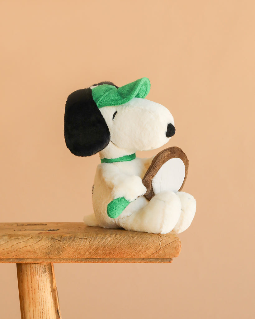 The Steiff Snoopy Tennis Player plush features Snoopy in a green visor holding a brown tennis racket, seated on a wooden stool against a beige background—perfect for Steiff collectors and Peanuts fans.