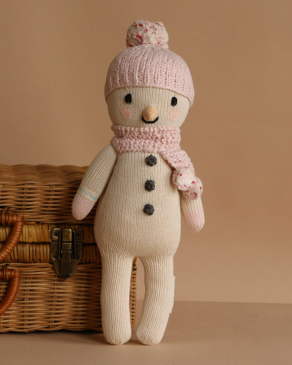 Cuddle + Kind Holly the Snowman is a knitted cotton doll with black button eyes, a beige carrot nose, and three black buttons. It wears a pink pom-pom hat and scarf, and sits by a wicker basket—ideal for handmade doll collections.