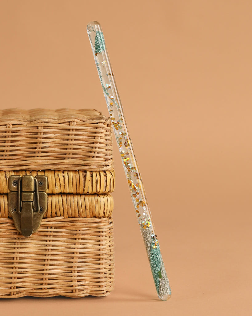 A Konges Sloejd Magical Glitter Wand - Green Mix leans against a closed woven wicker chest with a metal latch, set against a beige background.
