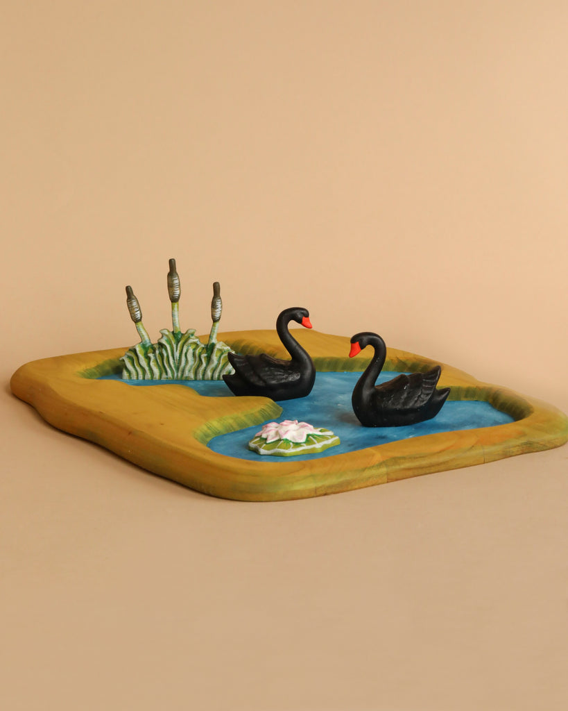 The Handmade Wooden Pond - Black Swans features two elegant black swan figurines gliding across a decorative pond adorned with lily pads and reeds, all set on a rectangular wooden base against a beige background.