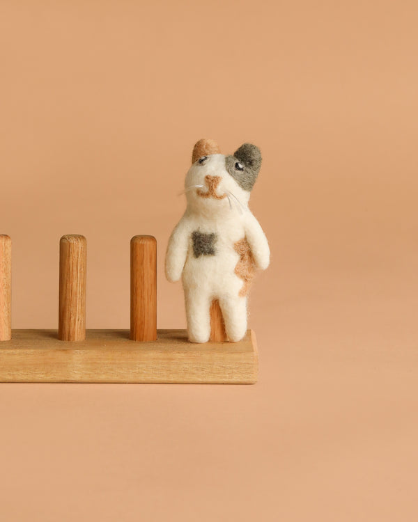 The Felt Finger Puppet: Calico Cat is a small, handmade wool figurine in brown, black, and white patches, standing on a light wooden base with four pegs. Crafted with care in Nepal.