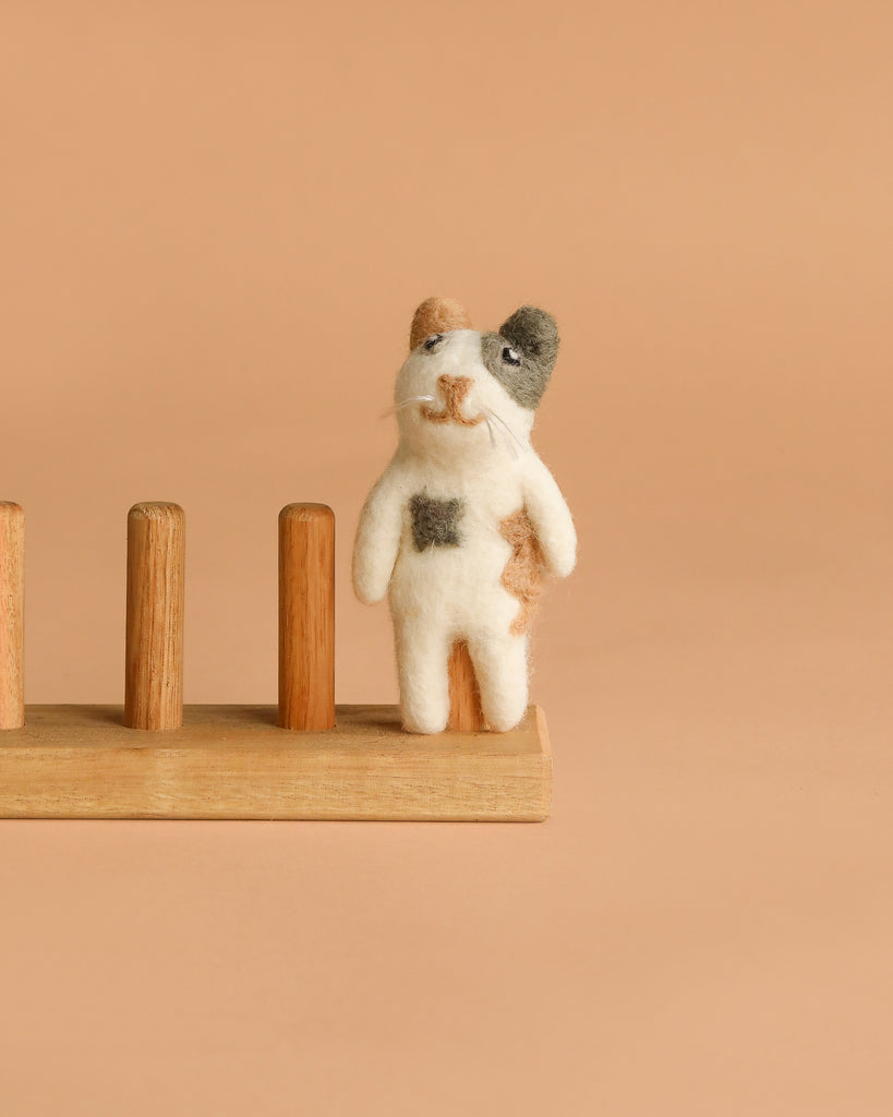 The Felt Finger Puppet: Calico Cat is a small, handmade wool figurine in brown, black, and white patches, standing on a light wooden base with four pegs. Crafted with care in Nepal.