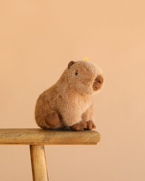 The Steiff Chip Capybara plush toy, featuring soft light brown fur, is shown seated on a wooden stool against a plain beige background.
