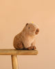 The Steiff Chip Capybara plush toy, featuring soft light brown fur, is shown seated on a wooden stool against a plain beige background.