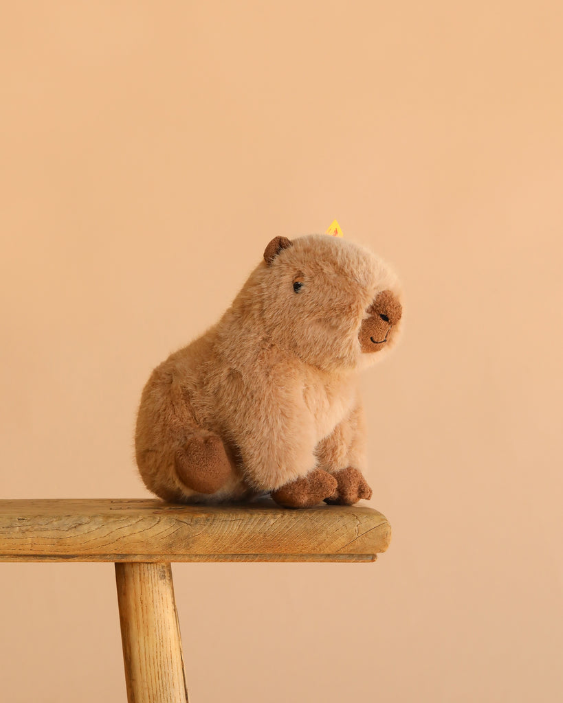 The Steiff Chip Capybara plush toy, featuring soft light brown fur, is shown seated on a wooden stool against a plain beige background.