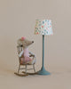 A Maileg Big Sister Set - Gift Wrapped mouse wearing a striped outfit and a pink hat sits on a miniature rocking chair next to a table lamp with a floral shade.