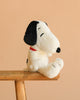 The Steiff Peanuts Snoopy plush, featuring black ears and a red collar, is displayed on a wooden stool against a plain beige background.