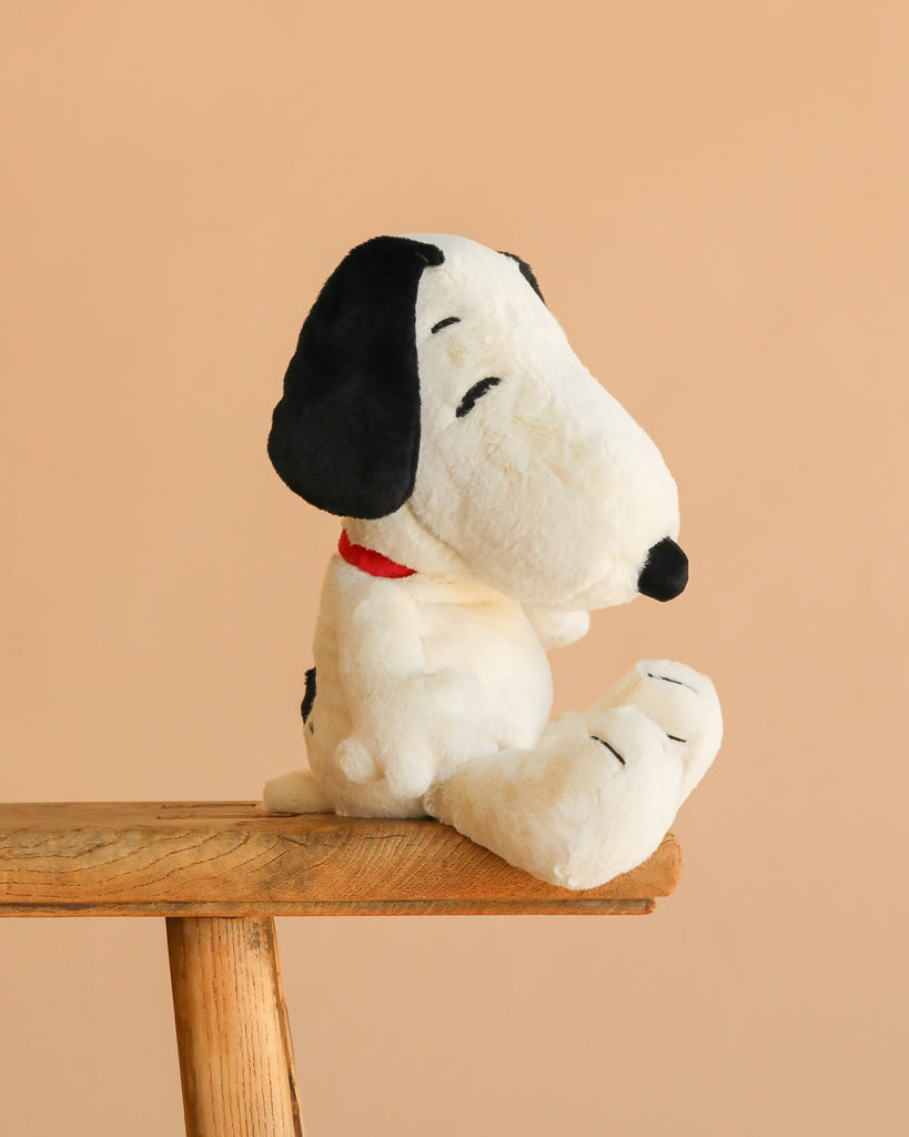 The Steiff Peanuts Snoopy plush, featuring black ears and a red collar, is displayed on a wooden stool against a plain beige background.