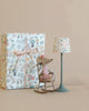 A Maileg Big Sister Set - Gift Wrapped sits on a rocking chair, facing a large floral-print gift box next to a miniature floor lamp with a matching floral shade, set against a soft beige background.