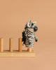 The Felt Finger Puppet: Zebra, a small striped animal made of felt, is displayed on a wooden stand against a plain beige background.