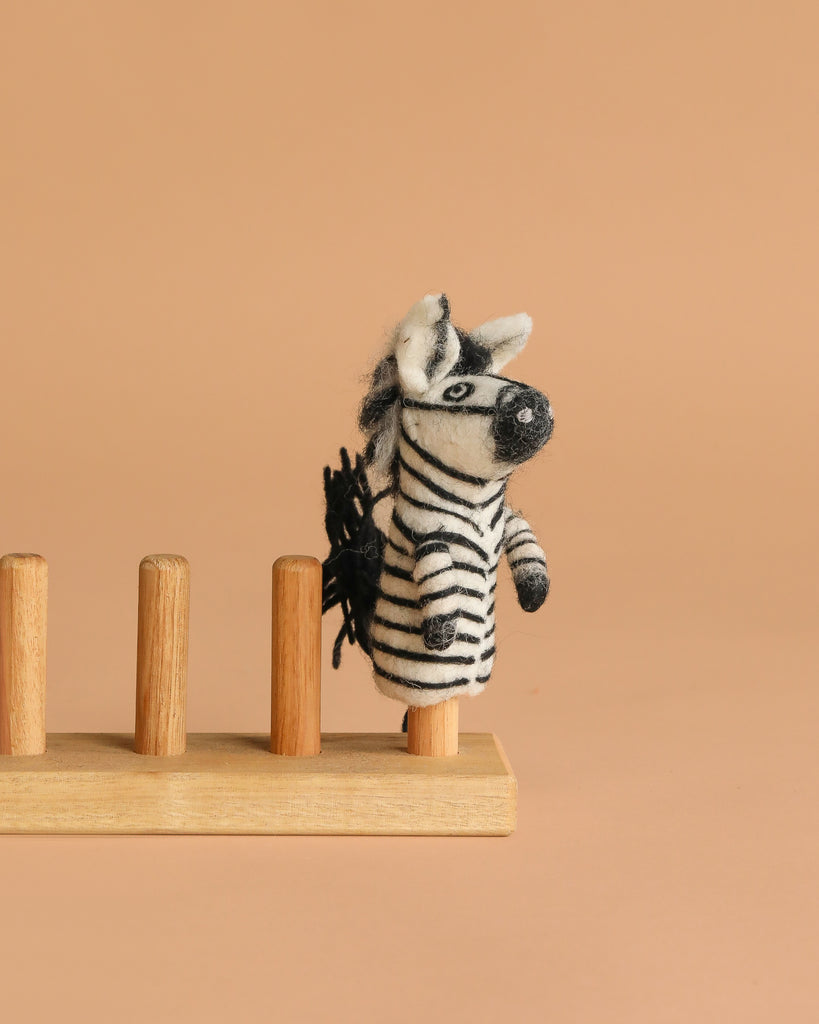 The Felt Finger Puppet: Zebra, a small striped animal made of felt, is displayed on a wooden stand against a plain beige background.