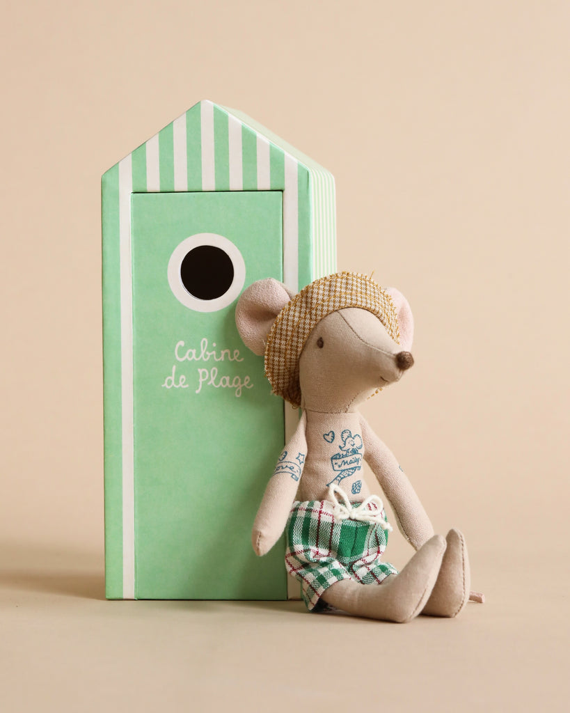 A Maileg Beach Mouse - Dad in swim trunks and a straw hat sits beside a green striped beach house labeled "cabine de plage." The background is a plain beige color.