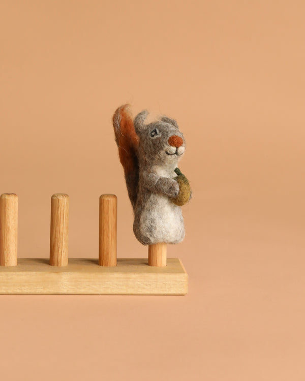 The Felt Finger Puppet: Gray Squirrel, an adorable woodland gift for kids, holds an acorn and stands on one of four wooden pegs attached to a simple base, displayed against a plain beige background.