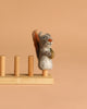 The Felt Finger Puppet: Gray Squirrel, an adorable woodland gift for kids, holds an acorn and stands on one of four wooden pegs attached to a simple base, displayed against a plain beige background.
