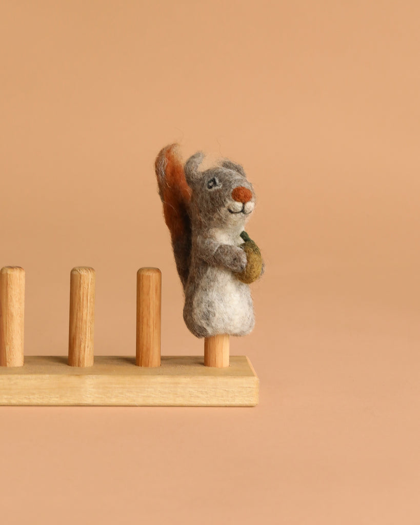 The Felt Finger Puppet: Gray Squirrel, an adorable woodland gift for kids, holds an acorn and stands on one of four wooden pegs attached to a simple base, displayed against a plain beige background.