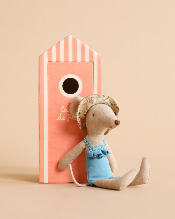 A Maileg Beach Mouse - Mom wearing a blue swimsuit and a floral hat sits with legs extended in front of a coral-colored beach house with white stripes on the roof and a circular opening. The background is a plain, light beige color, perfect for vacation time.