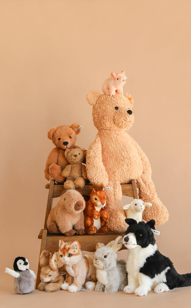 The Steiff XL Jimmy Teddy Bear, 22", sits on a wooden step stool, surrounded by assorted plush animals—dog, bunny, penguin, piglet, and fox—some with the iconic Steiff Button in Ear, all set against a beige background.