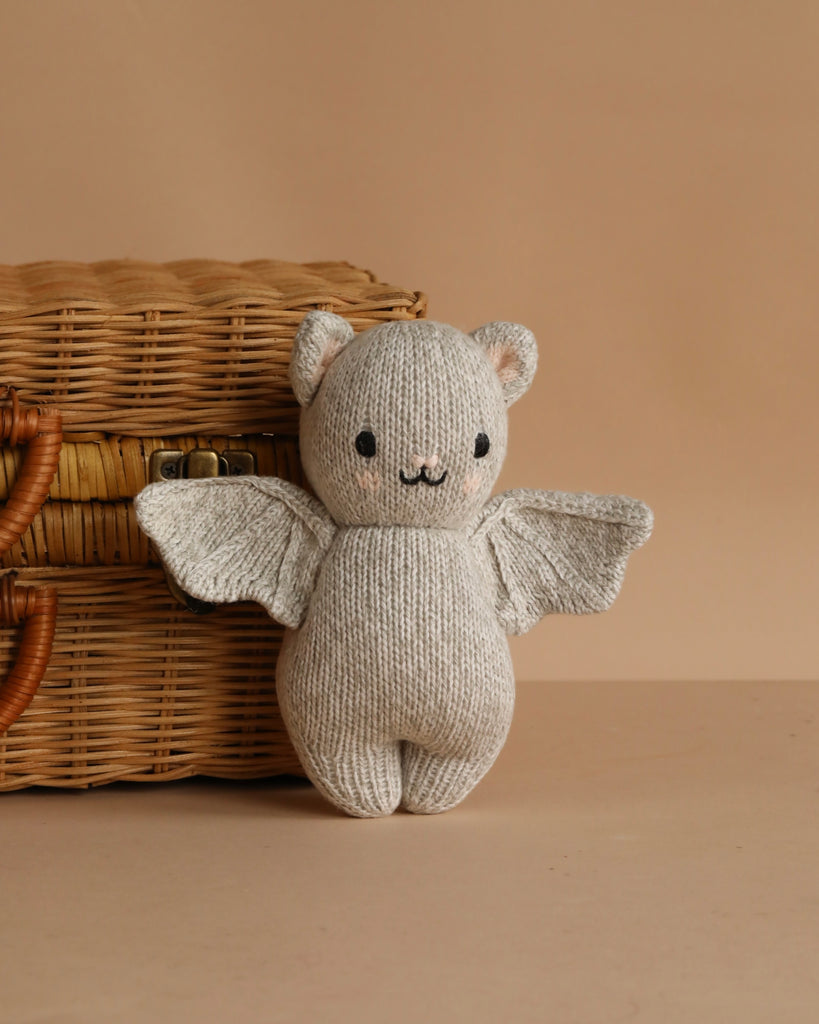 The Cuddle + Kind Baby Limited Edition Grey Bat, a light gray plush toy with bat wings, round ears, and a smiling face made from heirloom-quality Peruvian cotton yarn, sits before a wicker basket on a beige surface.