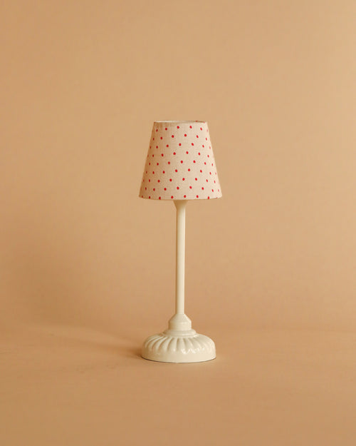 A miniature, white table lamp with a patterned shade adorned with small red dots against a muted beige backdrop provides gentle lighting. Its design is evocative of the Maileg Mouse Size Vintage Floor Lamp - Sand (With Battery), bringing charm and powered by an efficient coin cell battery (CR1632).