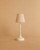 A miniature, white table lamp with a patterned shade adorned with small red dots against a muted beige backdrop provides gentle lighting. Its design is evocative of the Maileg Mouse Size Vintage Floor Lamp - Sand (With Battery), bringing charm and powered by an efficient coin cell battery (CR1632).