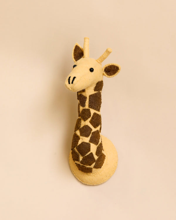 A handcrafted felt giraffe wall decor - mini with brown spots and a smiling face stands against a plain, light beige background.