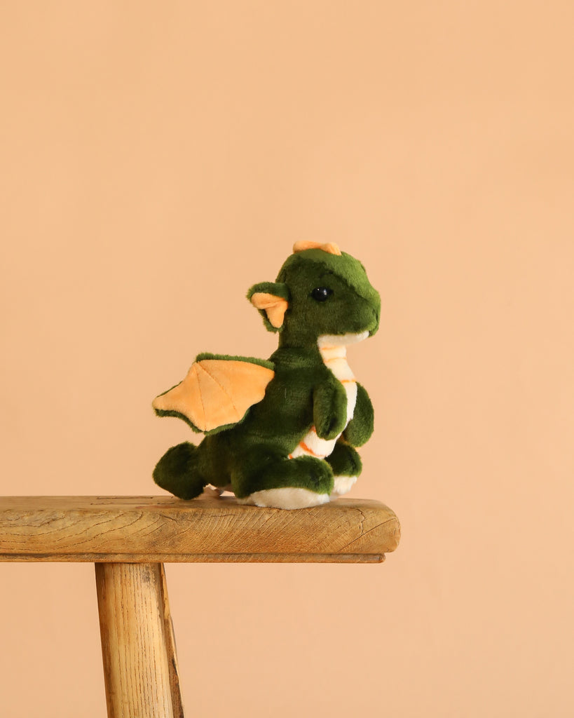 The Steiff Raudi Green Dragon Boy plush toy sits on a wooden stool against a plain peach-colored background.