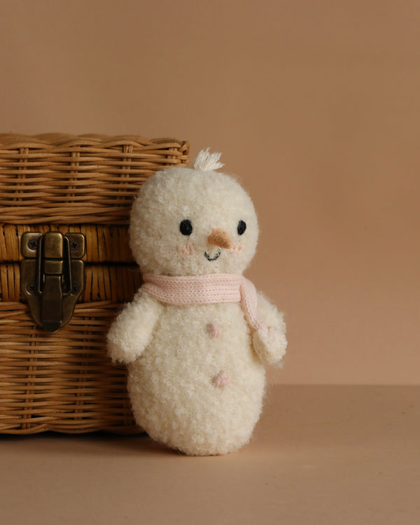 The Cuddle + Kind Baby Snowman - Blush, a small fair trade plush toy with black eyes, a pink scarf, and pink buttons, stands beside a closed wicker basket against a beige background.
