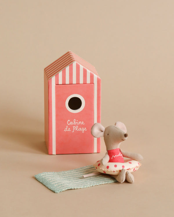 A Maileg Beach Mouse - Little Sister in a polka dot dress sits next to a pink striped beach house labeled "cabine de plage" on a beige background.