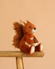 A Steiff Phil Squirrel, 8", with a Button in Ear and fluffy tail, sits on a wooden bench holding a small fabric acorn against a plain beige background.