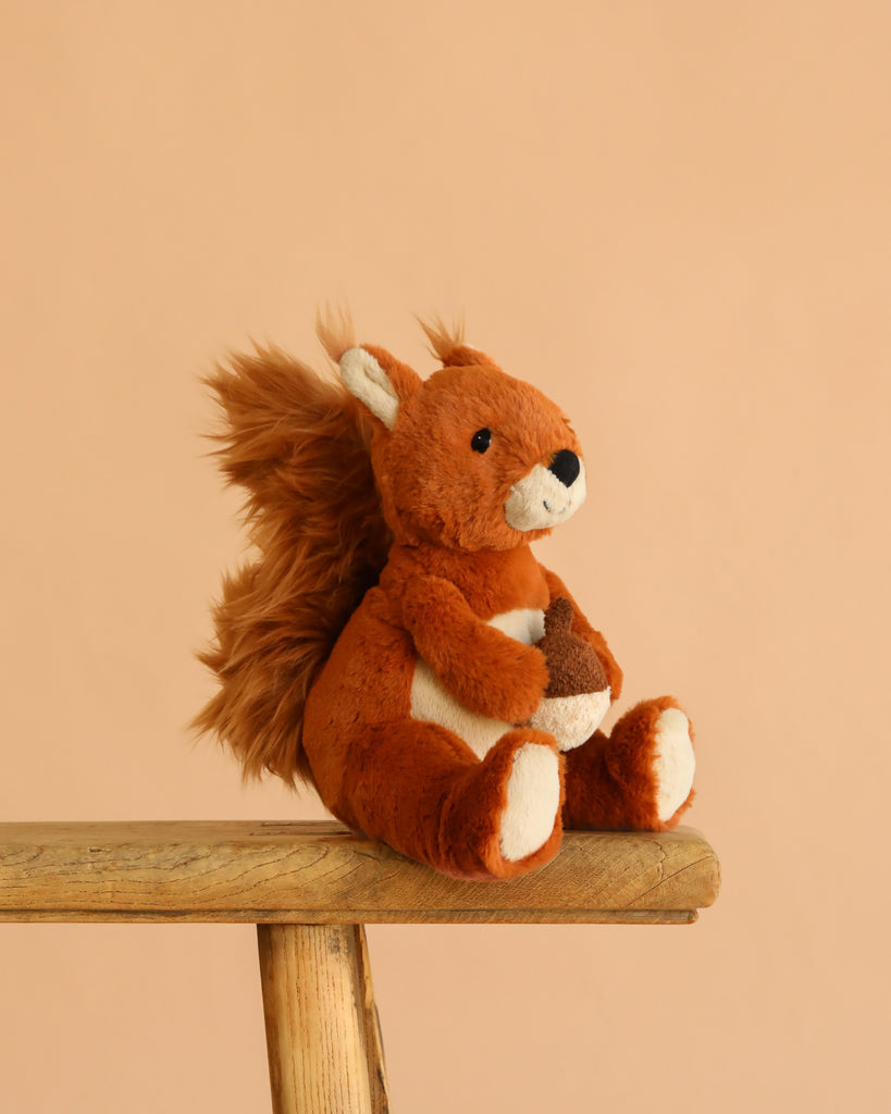 A Steiff Phil Squirrel, 8", with a Button in Ear and fluffy tail, sits on a wooden bench holding a small fabric acorn against a plain beige background.