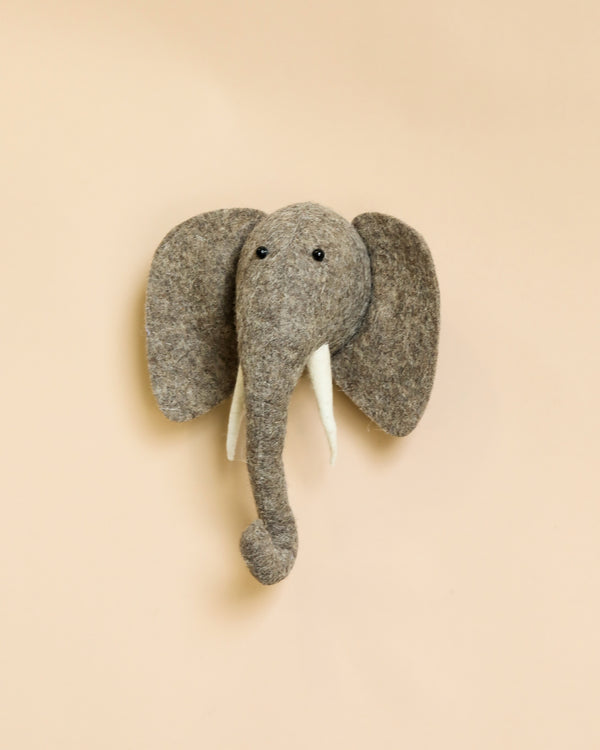 A Handcrafted Felt Elephant with Tusks Wall Decor - Mini mounted on a beige background, featuring large grey ears, a long trunk, and white tusks.