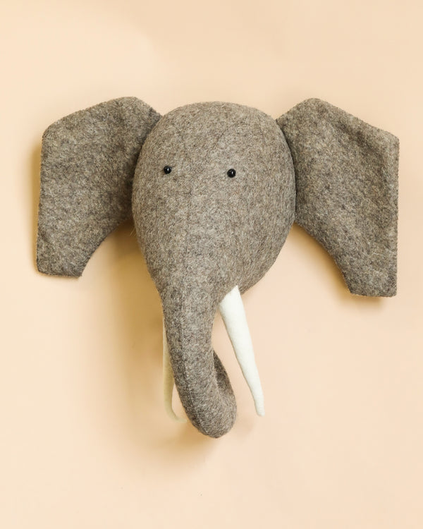 A handcrafted Felt Elephant with Tusks wall mount with large ears and tusks against a light peach background.